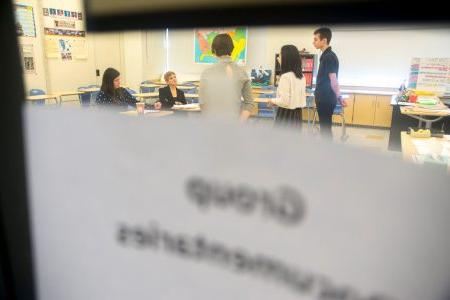 In the foreground is an out-of-focus paper sign on a door's glass window, while inside the room several students stand and speak to a team of judges.