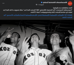 A screenshot of a Facebook post. There is text at the top and an image on the bottom. The image is a black and white photograph of three baseball players from a view around their waists looking up at them. The three men are smiling and looking forward and not at the camera. Two are wearing Red Sox jersey’s, one is bare-chested. They each hold up their index and middle fingers, showing the back of their hands to the camera, the one on the very right is holding a baseball in the same hand. There is a microphone held up below them, close to the camera, and the background has a ceiling and pipes. 