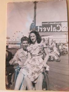 Photograph of Sammy in his uniform in a wheelchair on the boardwalk with a ferris wheel in the background. Bernice is in a dress and sitting on his lap.