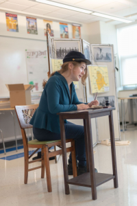 1 student in a classroom, sitting in a chair at a small desk with a pen in hand, leaning forward, enacting a performance 