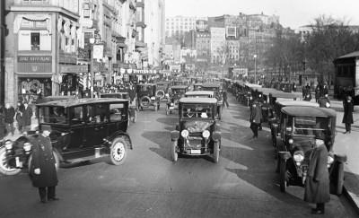 Black and white photograph depicting a street crowded with vehicles. Two men can be seen in the foreground directing traffic.