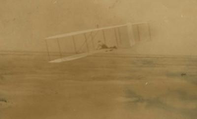 Yellowed photograph showing a glider with two wings and a person lying on the middle of it while in mid-flight