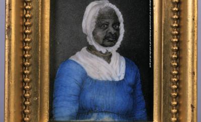 A framed portrait of a Black woman wearing a blue dress with drawstrings at the neck and waist, a white fichu tucked into her dress at the neck, a white cap, and a gold beaded necklace.