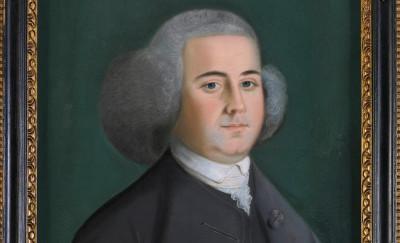 portrait of a white man wearing a gray wig, unbuttoned gray coat, gray vest, and white collar with arms behind his back against a dark background. the portrait is bordered by a black and gold frame, with a gold plaque labeled "John Adams"